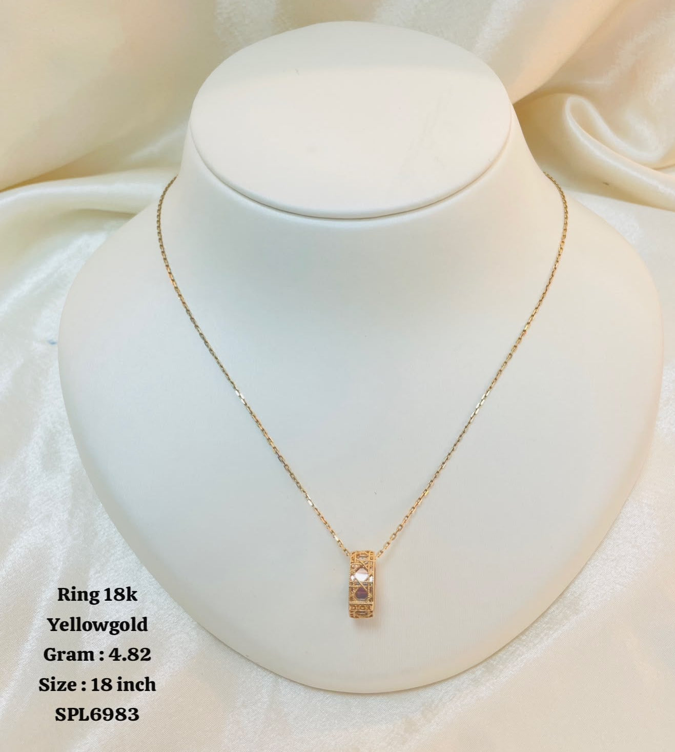 A delicate yellow gold chain necklace with a small rectangular pendant displayed on a white jewelry bust. The background is a soft, draped white fabric.