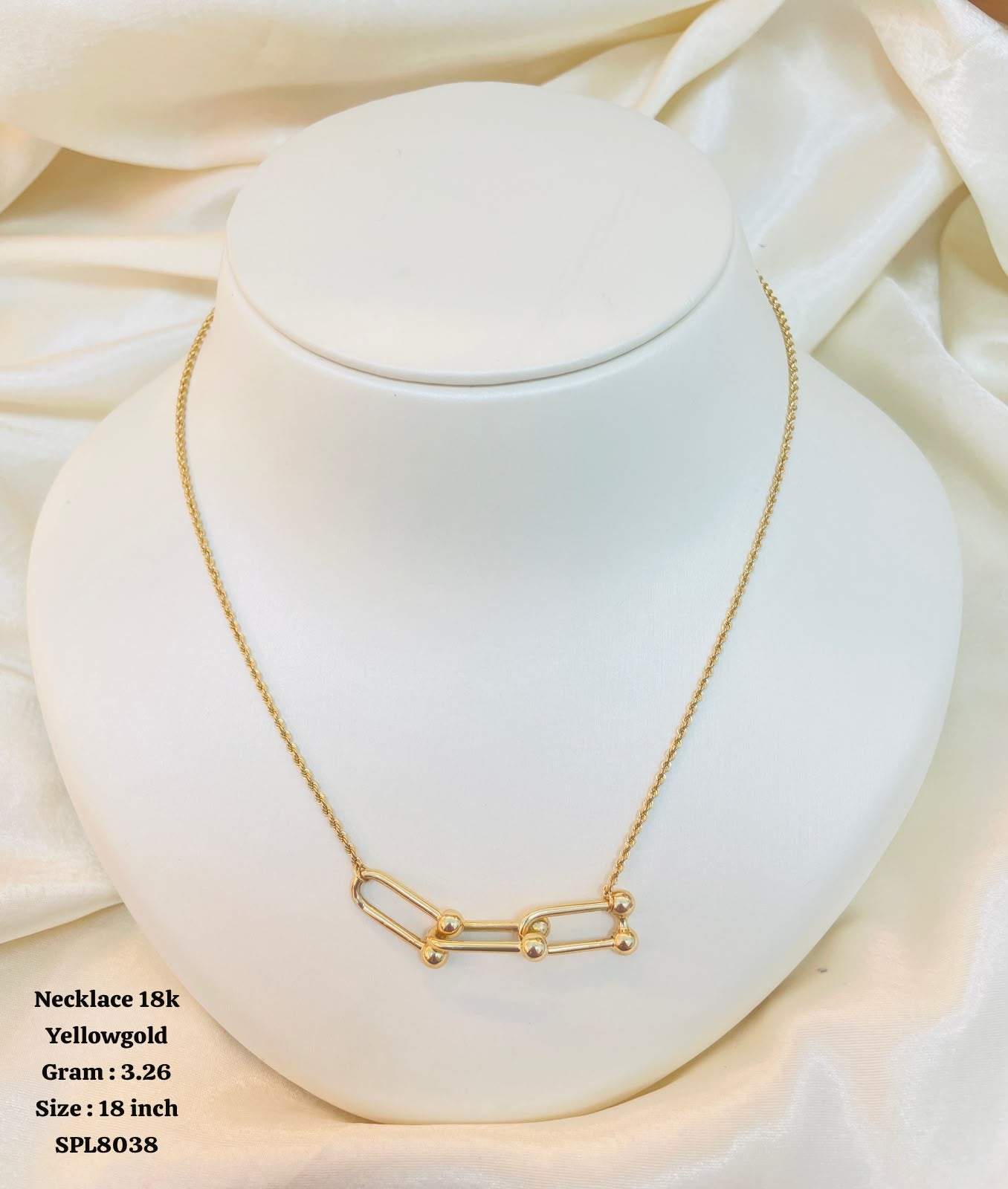 A gold necklace displayed on a white jewelry bust against a soft cream satin background. The necklace has a delicate chain and a pendant made of linked oval shapes with small bead details.