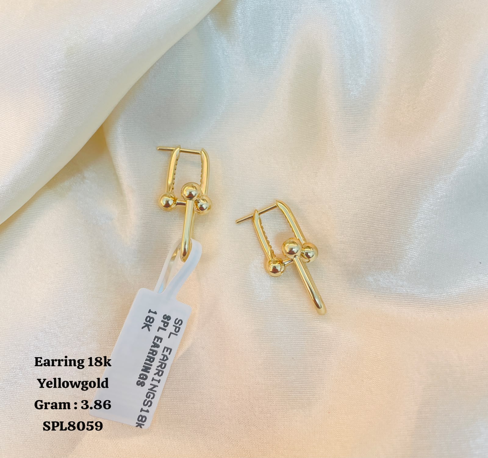 A pair of gold huggie hoop earrings with small bead details, displayed on draped cream satin fabric with a white product tag attached. Text overlay on the bottom left reads 'Earring 18k Yellowgold Gram : 3.