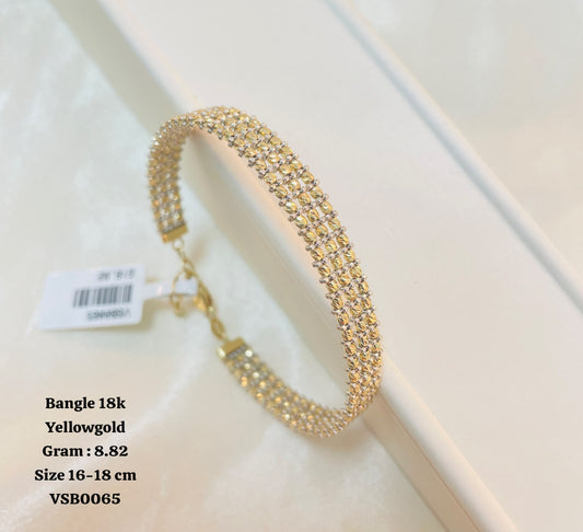 A yellow gold bangle bracelet with three rows of sparkling clear stones, displayed on a white textured surface with a product tag attached. Text on the left specifies the item as 'Bangle 18k Yellowgold' with weight and size details.